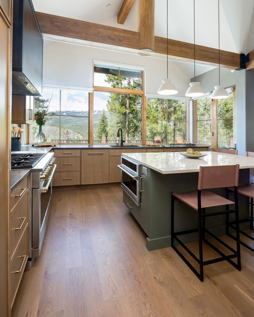 Modern mountain kitchen with natural wood cabinets, large center island with seating, stainless steel appliances, and expansive windows framing views of pine trees and snow-capped peaks.